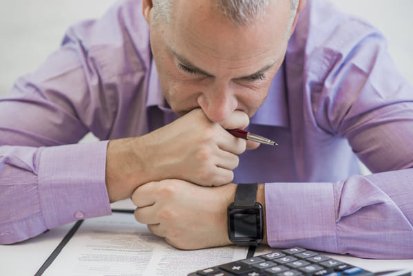 Homem de negócios de meia-idade com expressão preocupada, vestindo camisa social lilás, sentado à mesa com documentos e calculadora.