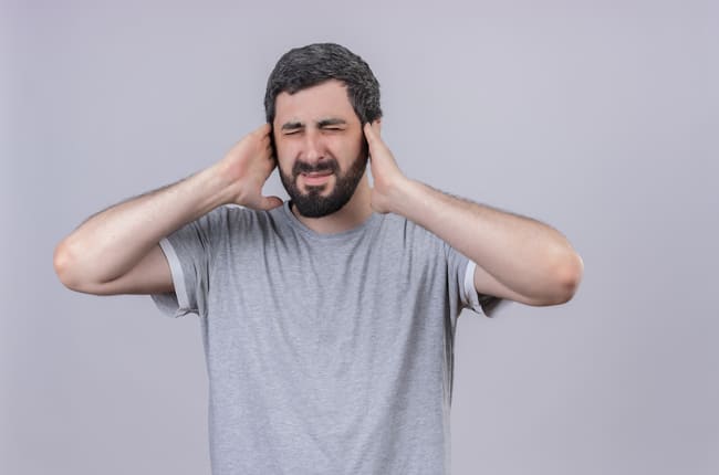 Homem com expressão de incômodo, olhos fechados e mãos tapando os ouvidos, representando desconforto ou irritação.